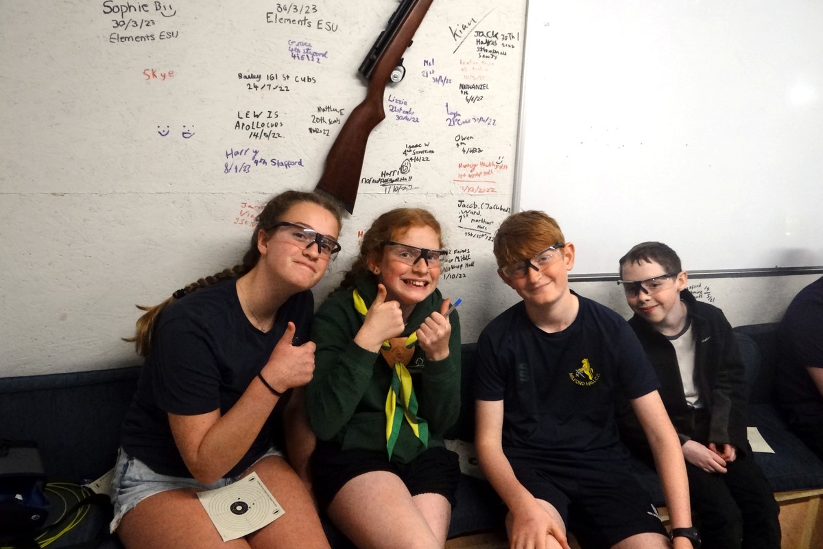 Image of four scout smiling, sat on a bench in an air rifle range